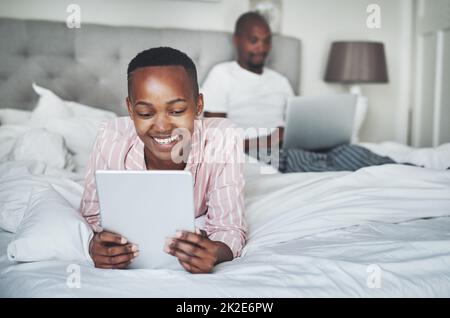 Turning on the day with their favourite devices. Shot of a happy young woman using a digital tablet while her husband uses a laptop in bed at home. Stock Photo