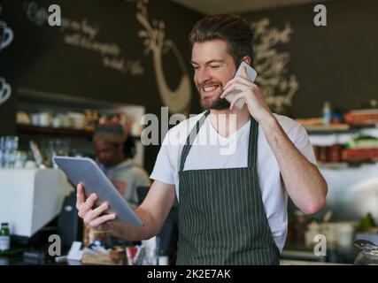 Just calling to let you know your order is ready. Shot of a young barista taking orders via cellphone and logging them on his tablet. Stock Photo