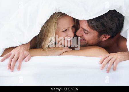 Keeping cosy. A couple lying under a duvet cover and cuddling affectionately while looking into each others eyes. Stock Photo