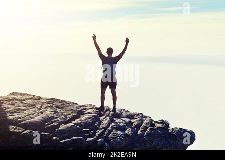 The best view comes after the hardest climb. Rearview shot of an unidentifiable young man raising his arms in triumph on a mountaintop. Stock Photo