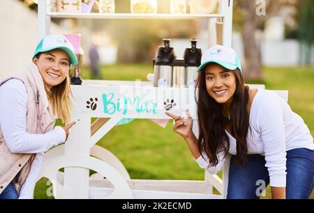 Come treat your sweet tooth. Portrait of two cheerful young women standing outside together next to their baked goods stall while looking at the camera outside. Stock Photo