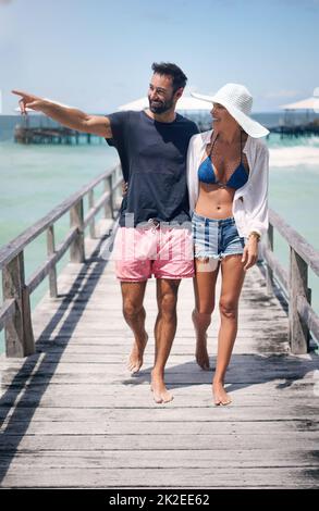 Lets go over there. Full length shot of a happy young couple walking arm in arm down a boardwalk during a vacation together. Stock Photo