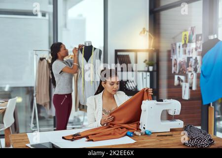 Making sure the fabric is of the highest quality. Shot of a young fashion designer sewing garments while a colleague works on a mannequin in the background. Stock Photo