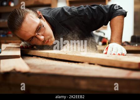 Looking great. Cropped shot of a focused young male carpenter blowing dust off of a piece of wood after sanding it inside of his workshop during the night. Stock Photo