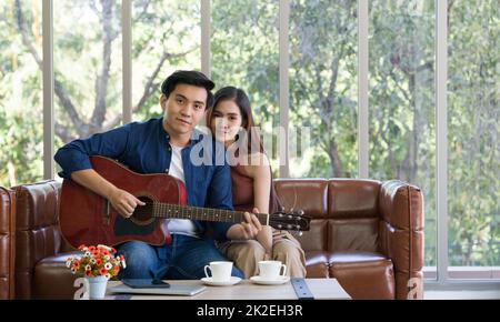 Young lovers spend time together on holidays in the living room. The young man wears comfortable clothes, play a guitar. Stock Photo