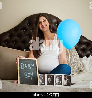 So much joy in one little boy. Portrait of a happy pregnant woman posing on her bed with an ultrasound picture, chalkboard and blue balloon. Stock Photo