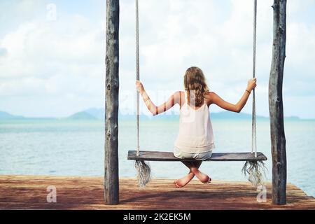 Spending the day in paradise. Shot of a young woman sitting on a swing overlooking the ocean. Stock Photo