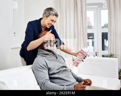 She never grows tired of surprising me. Shot of an attractive senior woman surprising her husband with a gift at home. Stock Photo