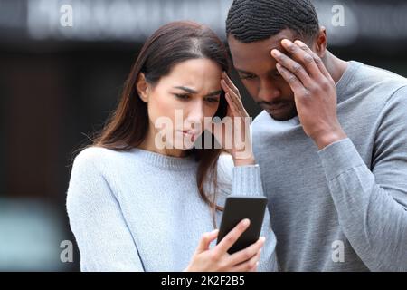 Worried interracial couple checking phone in the street Stock Photo
