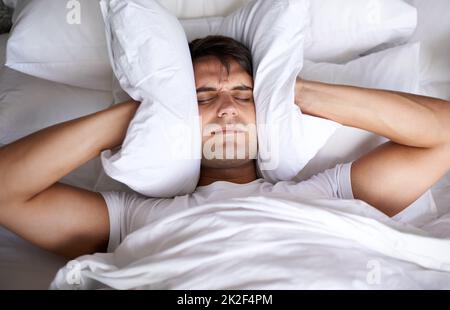 Whats with all the noise. a young man covering his ears with a pillow while trying to sleep. Stock Photo