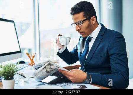 Commencing with his morning routine. Shot of a focused young businessman seated at his desk while drinking coffee and reading a newspaper in the office. Stock Photo