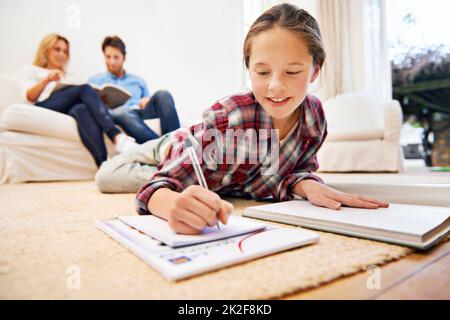 Homework done equals relaxing time. Shot of a little girl lying on the living room floor doing homework with her parents in the background. Stock Photo