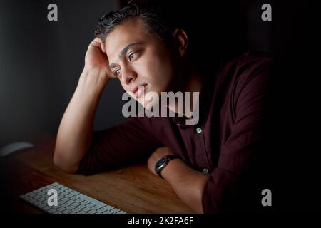 How long have I been staring at this screen now. Shot of a young businessman looking bored while working on a computer in an office at night. Stock Photo