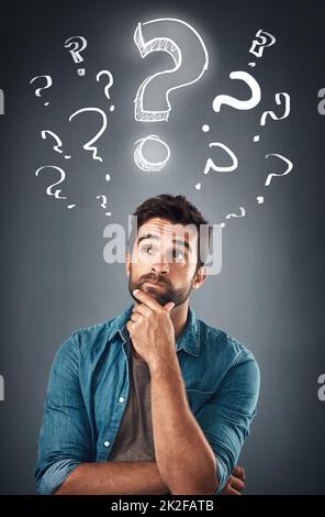 Hes full of bright ideas. Studio shot of a handsome young man looking thoughtful while standing underneath an illustration of question marks against a grey background. Stock Photo