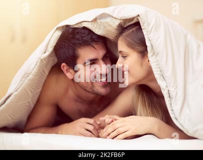 Love the one youre with. Shot of a loving young couple smiling at each other while lying under a blanket together. Stock Photo