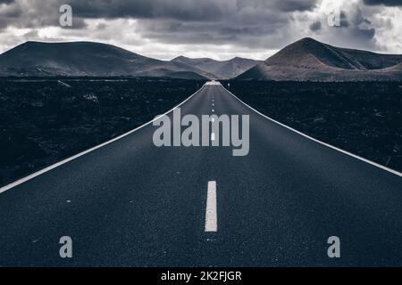 Endless road on a volcano in Timanfaya National Park in Lanzarote in the Canary Islands with a continuous line, black volcanic rocks on the side and volcanoes in mist in background. Stock Photo