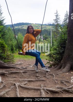 Woman with dark hair in a mustard sweater sitting on a swing and mountain view. Calm and quiet wanderlust concept moment when person feels happiness a Stock Photo