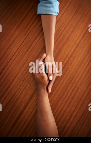 Reach out for some help. High angle shot of two unrecognizable peoples hands holding each other while resting on top of a wooden table. Stock Photo