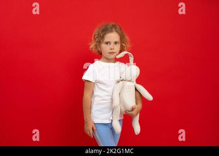 Charming little girl hold in hand favourite stuffed toy on empty red background, free copy space. Photo of nice child of kindergarten age with pretty Stock Photo
