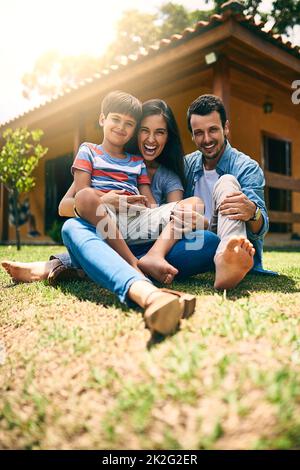 Doesnt matter where we are, as long as were together. Full length portrait of a happy young family of three sitting on their lawn in the backyard. Stock Photo