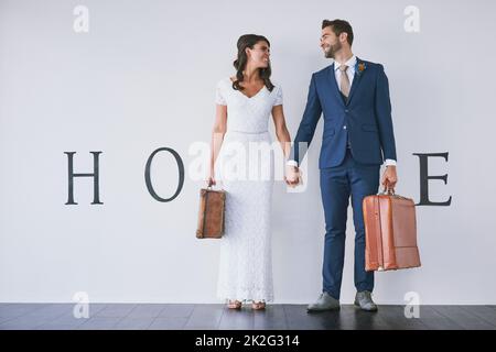 And so the adventure of marriage begins. Concept studio shot of a bride and groom making an M in the word home against a wall. Stock Photo