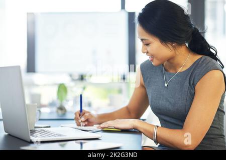 Putting plans into perspective and on paper. Cropped shot of a young businesswoman writing notes while working on a laptop in a modern office. Stock Photo
