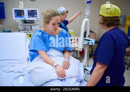 Preparing the final stages before surgery. Shot of a young pregnant woman sitting on her hospital bed and looking thoughtful with nurses in the background. Stock Photo