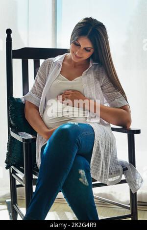 Counting the days to your arrival. Shot of a happy pregnant woman cradling her belly while relaxing in a chair at home. Stock Photo