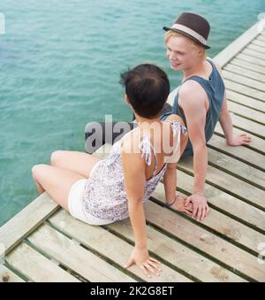 Sharing a quiet moment together. A high angle shot of a young couple talking while sitting on a dock with their legs hanging over the edge. Stock Photo