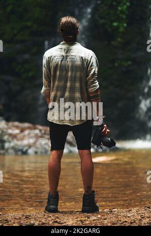 I want to capture photos of everything. Shot of a young man staring at a waterfall with his back turned and holding his camera. Stock Photo