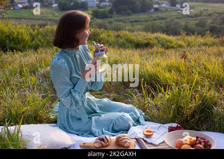 A woman in a long summer dress with short hair sitting on a white blanket with fruits and pastries and drinking lemonade. Concept of having picnic in Stock Photo