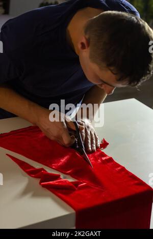 Hands of experienced worker in the handmade industry, performing cutting tasks on red fabric, on a white surface, the beginning of red hat production. Stock Photo