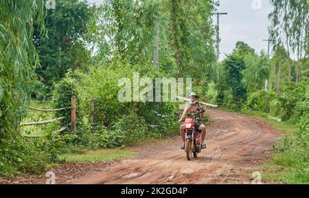 A farmer in a colourful shirt carries a bamboo tree trunk on his shoulder, while riding a motorcycle, taken at Sakon Nakhon, Thailand Stock Photo