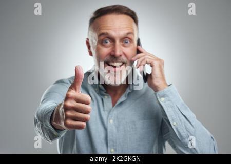 Weve been given the thumbs up to go ahead as planned. Studio portrait of a mature man showing thumbs up while talking on a cellphone against a grey background. Stock Photo