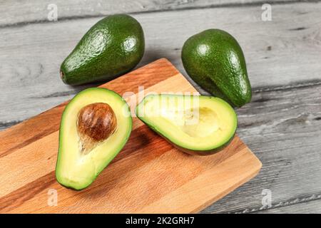 Avocado on wooden chopping board cut in half, seed visible, whole green avocado pears in background. Stock Photo