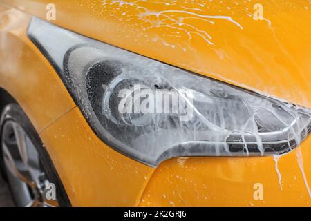 Front light of yellow car washed in carwash, jet water spraying, white shampoo and foam flowing on surface. Stock Photo