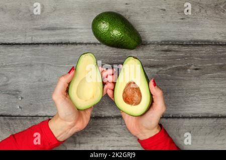 Tabletop view woman's hand holding two avocado halves, seed visible, whole avocado pear next to it. Stock Photo