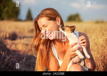 Young woman holding Jack Russell terrier puppy on her hand, trying to pose, but the dog is licking and chewing her ear and hair. Stock Photo