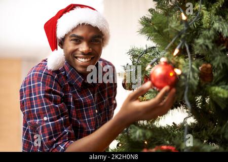 Christmas is my favourite time of year. a handsome young man wearing a santa hat decorating a christmas tree. Stock Photo