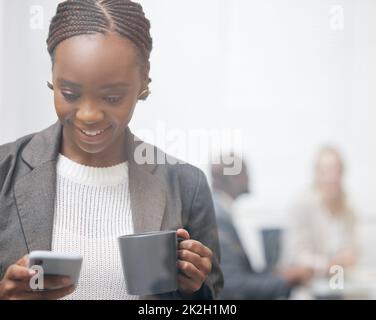 Shes always updated with whats new. Shot of a young businesswoman drinking coffee while using a cellphone in an office with her colleagues in the background. Stock Photo
