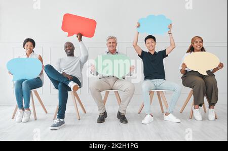 We want our opinions to be heard as well. Portrait of a diverse group of people holding colourful speech bubbles while sitting in line against a white background. Stock Photo