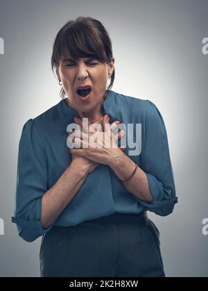 That really breaks my heart to hear. Studio shot of a senior woman holding her chest in pain against a grey background. Stock Photo