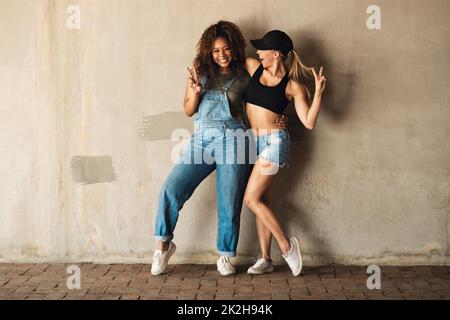 We know how to have fun. Portrait of two cheerful young women posing for for a photo while leaning against a wall outside during the day. Stock Photo
