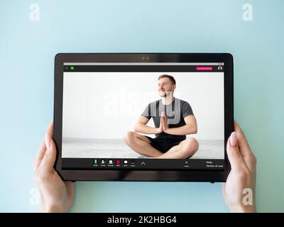 Yoga video class. Online meditation. Virtual training. Woman hands holding tablet with relaxed man in lotus pose on screen isolated on blue. Stock Photo