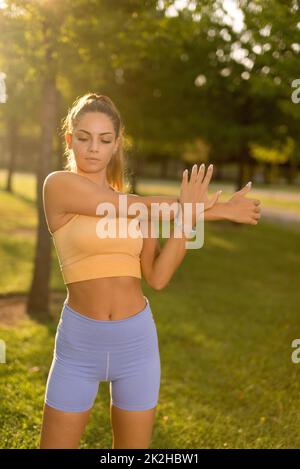 A woman with a yellow top and blue tights stretching arms and warming up in a public park in the morning. Stock Photo