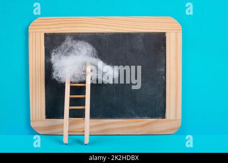 Ladder with a cloud on top, blank chalkboard, blue colored background, copy space, dreaming of success, creative and business concept, achieving new goals Stock Photo