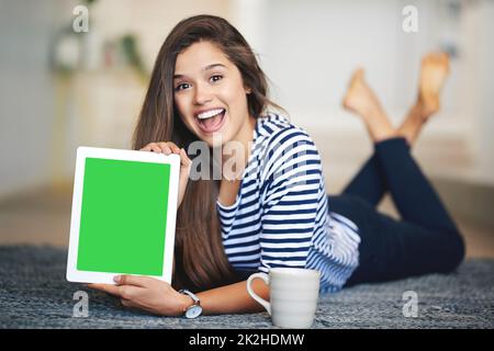 This is a site youre going to want to visit. Portrait of an ecstatic young woman lying on the floor at home holding up a digital tablet with a chroma key screen. Stock Photo