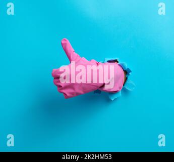 hand in a pink household rubber glove sticks out of the torn hole and shows a gesture okay, thumb up. Blue background Stock Photo