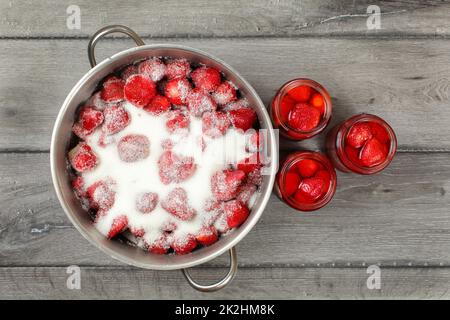 Tabletop view - steel put with strawberries covered in crystal sugar, three bottles with pickled strawberry next to it. Home made jam / compote preparation Stock Photo