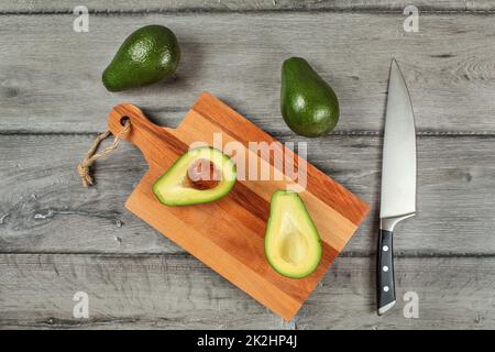 Tabletop view, avocado cut in half, seed visible, on a chopping board, chef knife, and two whole green pears next to in. Stock Photo